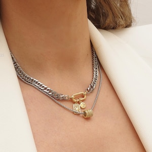 May include: A layered necklace featuring a silver curb chain and a delicate chain with a gold-tone pendant. The necklace includes a gold-tone clasp and a small, gold-tone charm with a geometric design and a small, gold-tone cylinder pendant.