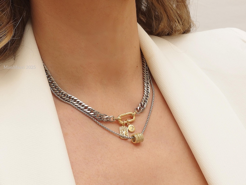 May include: A layered necklace featuring a silver curb chain and a delicate chain with a gold-tone pendant. The necklace includes a gold-tone clasp and a small, gold-tone charm with a geometric design and a small, gold-tone cylinder pendant.