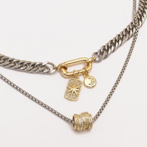 May include: A layered necklace with a silver curb chain and a delicate silver chain. The necklace features a gold-colored clasp and charms, including a starburst and a crescent moon, both adorned with small, clear stones.