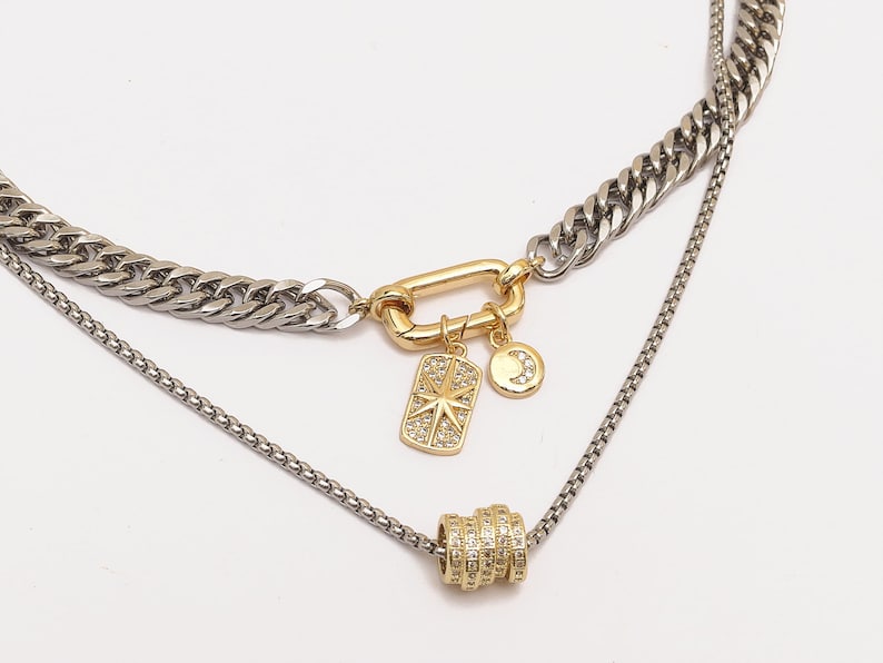 May include: A layered necklace with a silver curb chain and a delicate silver chain. The necklace features a gold-colored clasp and charms, including a starburst and a crescent moon, both adorned with small, clear stones.