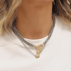 May include: A layered necklace featuring a thick silver chain and a thinner silver chain with a gold-colored clasp and pendant. The pendant has a small, square, gold-colored charm with a diamond-like pattern. The necklace is worn on a white shirt.
