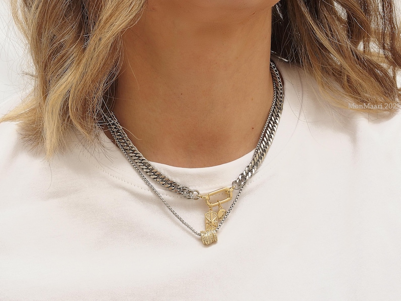 May include: A layered necklace featuring a thick silver chain and a thinner silver chain with a gold-colored clasp and pendant. The pendant has a small, square, gold-colored charm with a diamond-like pattern. The necklace is worn on a white shirt.