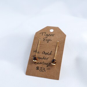 Puede incluir: Pendientes colgantes dorados con cuentas de ojo de tigre. Los pendientes se muestran en una etiqueta marrón con las palabras "Tigers Eye" y "14k Gold Threader earrings".