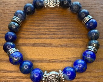 Pode incluir: Uma pulseira de contas com pedras esféricas azul escuro e azul marinho. Os detalhes metálicos prateados incluem espaçadores decorativos e duas contas focais maiores com desenhos de nós celtas. A pulseira é exibida em uma superfície de madeira.