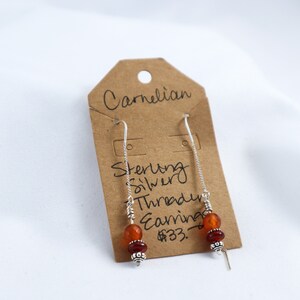 Puede incluir: Pendientes de hilo de plata de ley con cuentas de cornalina. Los pendientes tienen una cadena larga y delicada con un gancho en la parte superior y un grupo de cuentas naranjas y rojas en la parte inferior. Se muestran en una etiqueta marrón con la palabra "Carnelian".