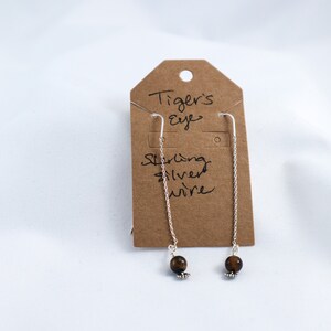 Puede incluir: Pendientes colgantes con cuentas de ojo de tigre y alambre de plata de ley. Se muestran en una etiqueta marrón con la inscripción "Tiger's Eye Sterling silver wire". Diseño sencillo y elegante.