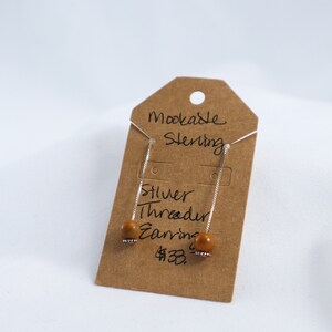 Puede incluir: Pendientes de hilo de plata con una pequeña cuenta redonda de color marrón claro en cada extremo. Los pendientes se muestran en una etiqueta marrón que dice "Mookable Sterling Silver Threader Earrings".