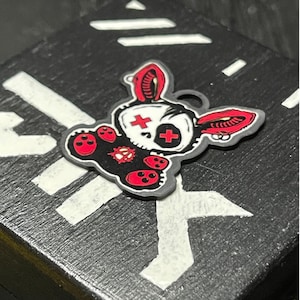 May include: A flat, black and white charm shaped like a stylized rabbit head with red accents. The rabbit features red cross-shaped eyes, red paw pads, and a red symbol on its chest. It has long ears with red inner details and small silver hoop earrings.
