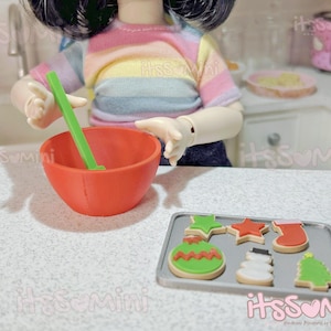 May include: A miniature kitchen scene with a doll holding a green spatula over an orange bowl. A baking sheet with Christmas-themed cookies is in front. The doll wears a rainbow striped shirt. The scene is set on a white countertop.