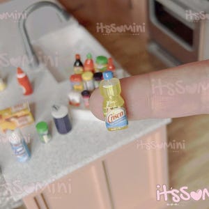 May include: Miniature yellow Crisco bottle with a blue and white label, held by a finger. Other miniature food items are visible in the background, including bottles, boxes, and containers, suggesting a kitchen or grocery store theme.