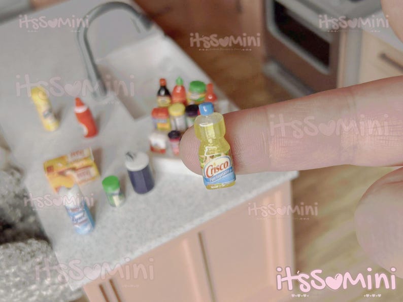 May include: Miniature yellow Crisco bottle with a blue and white label, held by a finger. Other miniature food items are visible in the background, including bottles, boxes, and containers, suggesting a kitchen or grocery store theme.