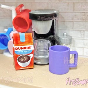 May include: Miniature kitchen scene with a coffee maker, a purple mug, and a Dunkin' French Vanilla coffee box. A red mug rack holds blue and red mugs. The coffee maker is black and clear, with a silver lid. The coffee box is orange and blue.