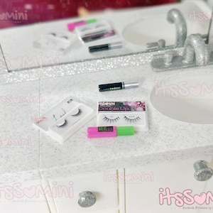 May include: Miniature vanity scene with beauty products. Includes a mirror, sink, and various makeup items like mascara and false eyelashes. The scene is set on a white countertop with glitter accents, creating a glamorous aesthetic.