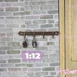 May include: Miniature bronze-colored key rack with four hooks, holding two keychains. The rack is mounted on a light gray brick wall, next to a wooden door. The text "1:12" is visible.