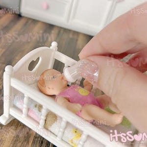 May include: A miniature white crib with a heart-shaped cutout, holding a doll dressed in a pink outfit. A small bottle is being held up to the doll's mouth. The scene is set on a wooden surface.