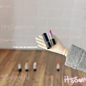 May include: Miniature black lipstick tube next to a silver and black lipstick with a bright pink lipstick. The text "LID CAN BE REMOVED" is visible. Several other miniature lipsticks are in the background.