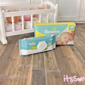 May include: A miniature white wooden crib with a baby doll inside, next to a package of Pampers diapers and a package of baby wipes. The Pampers package is yellow and teal, and the wipes package is teal and white.