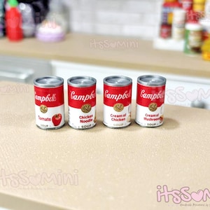 May include: Four miniature Campbell's soup cans with red and white labels. The cans are labeled Tomato, Chicken Noodle, Cream of Chicken, and Cream of Mushroom. The cans are on a light brown surface.
