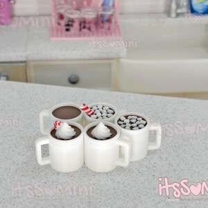 May include: Miniature white mugs filled with hot chocolate, whipped cream, and candies. Some mugs have peppermint sticks. The mugs are arranged on a speckled countertop, suggesting a cozy, dollhouse-themed scene.