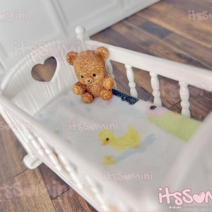 May include: A miniature white crib with a heart-shaped cutout on the headboard. A small, brown teddy bear sits inside the crib on a patterned blanket with a yellow duck design. The crib is a charming, decorative item.