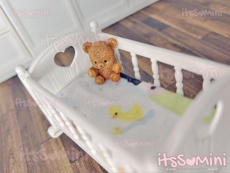 May include: A miniature white crib with a heart-shaped cutout on the headboard. A small, brown teddy bear sits inside the crib on a patterned blanket with a yellow duck design. The crib is a charming, decorative item.