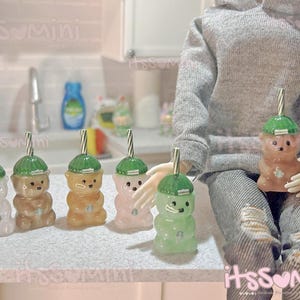 May include: A collection of miniature bear-shaped drink containers in various colors, including clear, brown, orange, pink, and green. Each container has a green lid and a striped straw. The containers are arranged on a white surface.