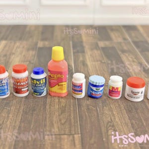 May include: A collection of miniature medicine bottles and jars, including brands like Pepcid, Advil, and Vicks VapoRub. The containers have various colors, including red, blue, white, and orange, and are arranged on a wooden surface.
