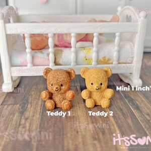 May include: Two miniature teddy bears, one light brown and one golden yellow, sit in front of a white doll-sized crib. The bears are labeled "Teddy 1" and "Teddy 2". The text "Mini 1 inch Teddy" is also visible.