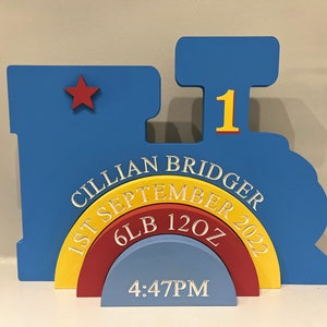 May include: A blue wooden train-shaped sign with a red star, the number 1, and the text "Cillian Bridger 1st September 2022 6lb 12oz 4:47PM".