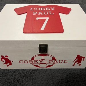 May include: White wooden box with a red soccer jersey on the lid. The jersey has the name "Coby Paul" and the number "7" on it. The box also has a soccer ball and two soccer players on the front.