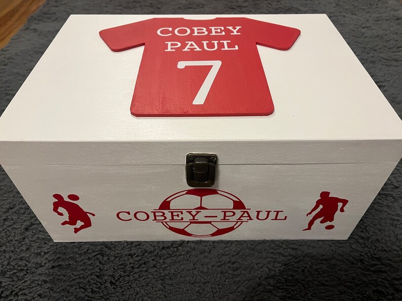 May include: White wooden box with a red soccer jersey on the lid. The jersey has the name "Coby Paul" and the number "7" on it. The box also has a soccer ball and two soccer players on the front.