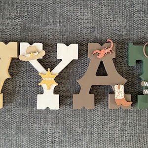 May include: Wooden letters spelling the name "WYATT" decorated with western-themed imagery, including a cowboy hat, sheriff's star, horseshoe, cactus, boot, scorpion, and a sign that reads "Wild West".