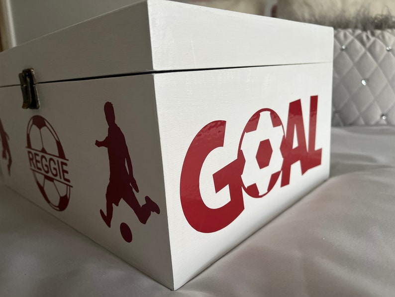May include: White wooden box with a red and white soccer ball design. The box has the word "GOAL" written in red letters and a soccer player silhouette. The soccer ball has the name "REGGIE" written on it.
