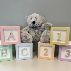 May include: A set of six wooden blocks with pastel colours and white letters and numbers. The blocks spell out "ABC" and "123". A grey teddy bear sits on top of the blocks.