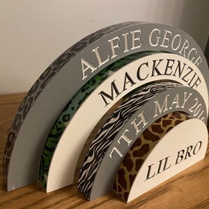 Puede incluir: Un arco de madera gris, blanco y negro con estampado de leopardo, estampado verde y blanco, estampado de cebra y estampado de jirafa. El arco est&aacute; grabado con los nombres "Alfie George Mackenzie", "7th May 201" y "Lil Bro".