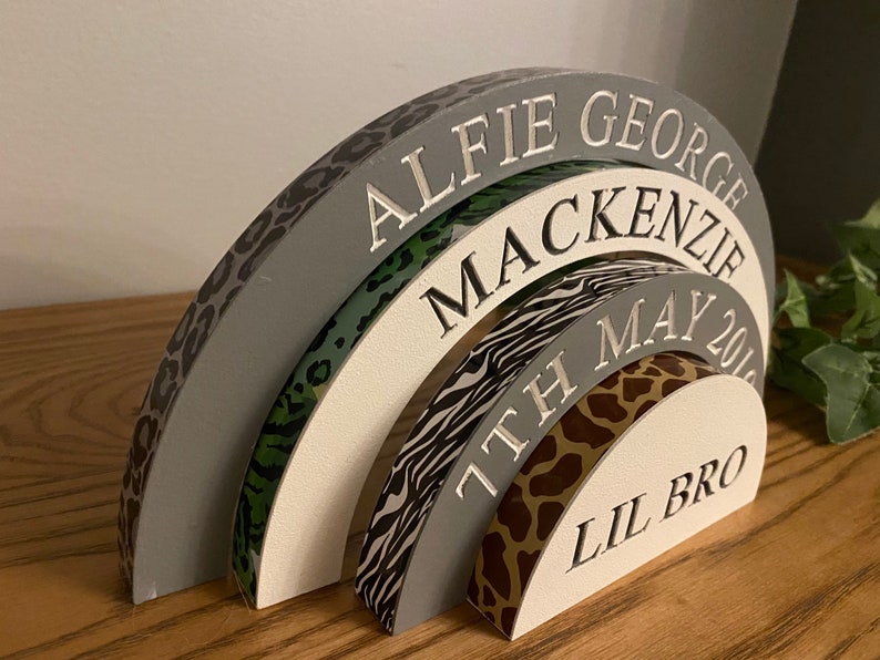 Puede incluir: Un arco de madera gris, blanco y negro con estampado de leopardo, estampado verde y blanco, estampado de cebra y estampado de jirafa. El arco est&aacute; grabado con los nombres "Alfie George Mackenzie", "7th May 201" y "Lil Bro".