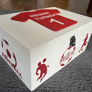 May include: White wooden box with a red football shirt on top. The shirt has the name "Reggie Turner" and the number "1" on it. The box has a red football design on the side with the text "Reggie" and "Football".