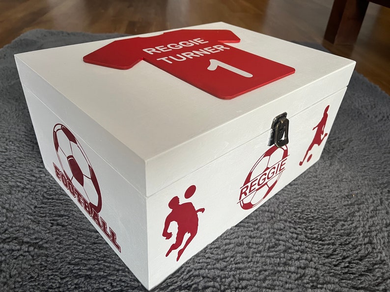 May include: White wooden box with a red soccer jersey on top. The jersey has the name "Reggie Turner" and the number "1" on it. The box has a red soccer ball design on the side with the text "Reggie" and "Football".
