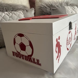 May include: White wooden box with a red lid and red football-themed designs. The box has a silver latch and the words "FOOTBALL" and "REGGIE" are printed on the side.