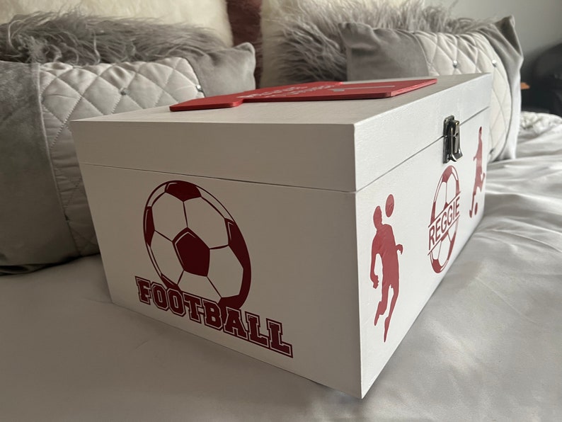 May include: White wooden box with a red lid and red football-themed designs. The box has a silver latch and the words "FOOTBALL" and "REGGIE" are printed on the side.