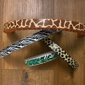 Puede incluir: Cuatro piezas de adorno de tela con diferentes patrones. Una pieza es marr&oacute;n con estampado de jirafa, otra es blanco y negro con estampado de cebra, otra es verde con estampado de camuflaje y otra es marr&oacute;n con estampado de leopardo.