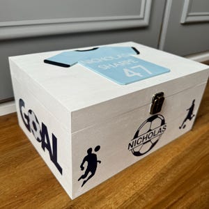 May include: White wooden box with a blue and black soccer jersey design on the top. The box has the word "GOAL" printed on the side in black and white. The box also has a black and white soccer player graphic and the name "Nicholas" printed on the side.