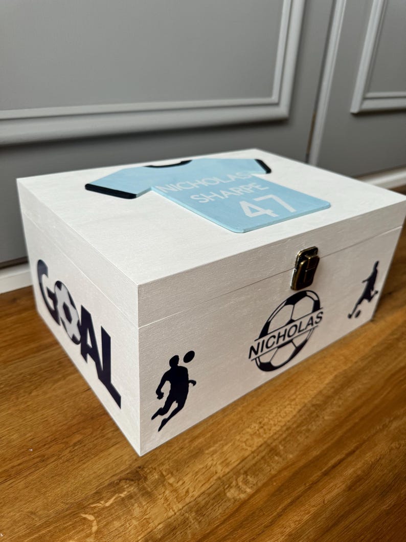 May include: White wooden box with a blue and black soccer jersey design on the top. The box has the word "GOAL" printed on the side in black and white. The box also has a black and white soccer player graphic and the name "Nicholas" printed on the side.