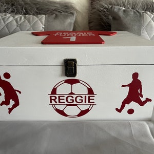 May include: White wooden keepsake box with a red soccer jersey design on the lid. The box has a red soccer ball design with the name "Reggie" on it. There are two red soccer player silhouettes on the box.