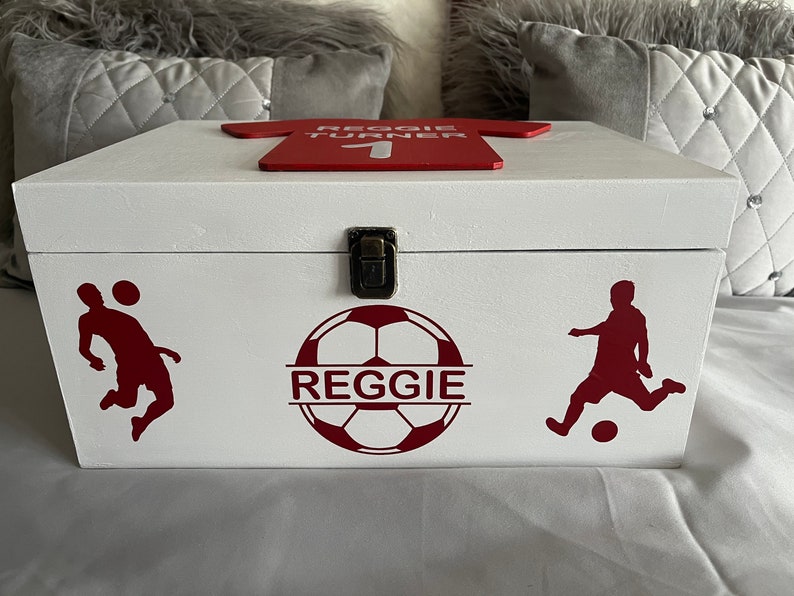 May include: White wooden keepsake box with a red soccer jersey design on the lid. The box has a red soccer ball design with the name "Reggie" on it. There are two red soccer player silhouettes on the box.