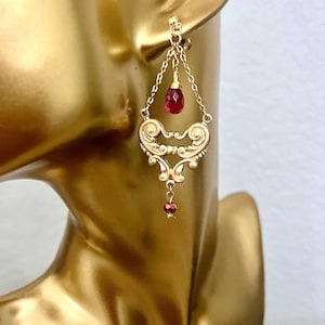 May include: Gold dangle earrings with a red gemstone pendant. The earrings feature a delicate, ornate design with a dangling chain and a small red gemstone at the bottom.