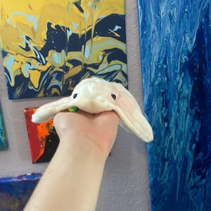 May include: A white plush stingray toy with black eyes. The toy is being held by a hand in front of a wall with colourful abstract paintings.