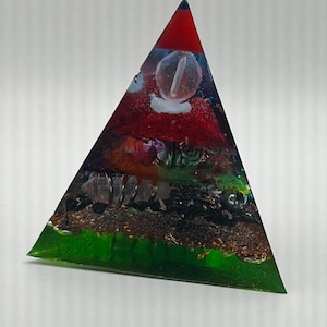 Puede incluir: Una pirámide de cristal de múltiples capas con un cristal de cuarzo transparente en la parte superior. La pirámide está hecha de resina verde, negra, roja y azul con motas doradas.