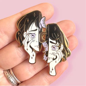 May include: Gold-tone enamel pin featuring two women's faces, each with a different expression. One face is sad with a tear, the other is smiling. Both faces are partially obscured by butterflies and a crescent moon.