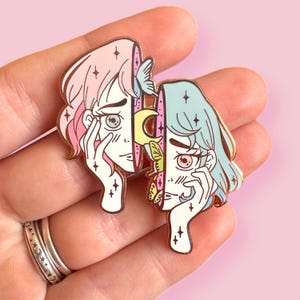 May include: Two enamel pins depicting stylized faces of women with pastel pink and blue hair. The faces are split in half, with one side showing a crescent moon and butterflies, and the other side showing stars. The pins are gold-toned.